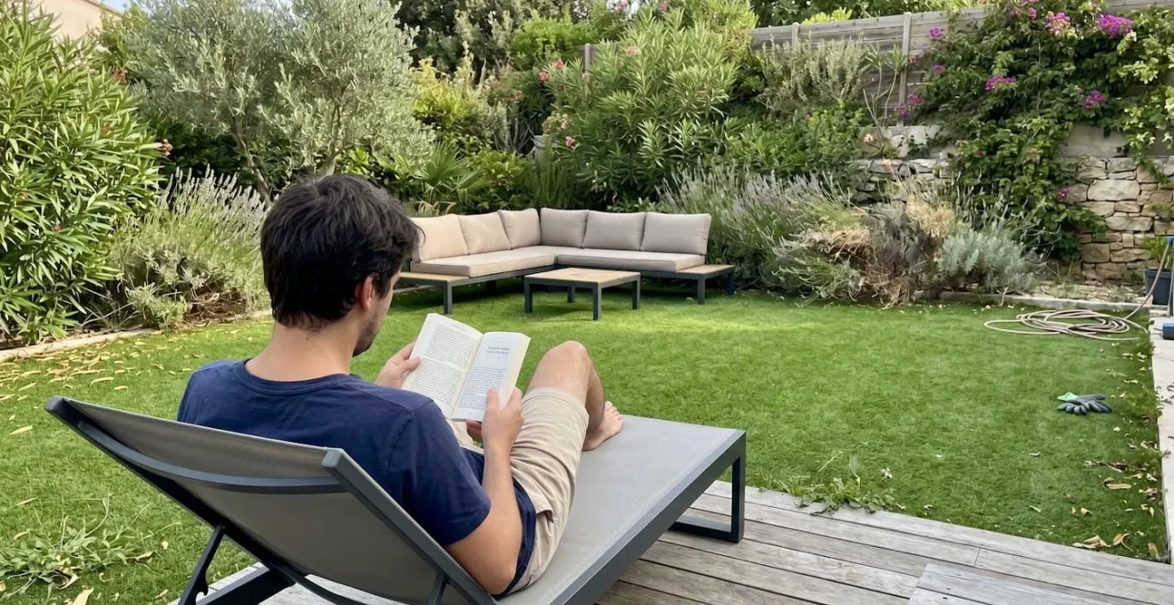 Coin détente de jardin aménagé avec pelouse synthétique verte, personne de dos assise sur transat, végétation méditerranéenne en arrière-plan