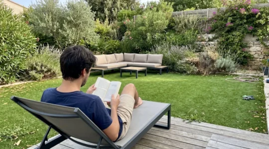 Coin détente de jardin aménagé avec pelouse synthétique verte, personne de dos assise sur transat, végétation méditerranéenne en arrière-plan